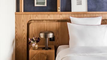 Una cama bien hecha con ropa blanca, una mesita de noche de madera con una lámpara moderna, y un jarrón con flores.