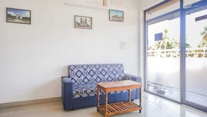 A room with a patterned sofa, a wooden table, and a large window with a view of a building and greenery. - La Bell Residency Near Auroville Beach (Vanur)