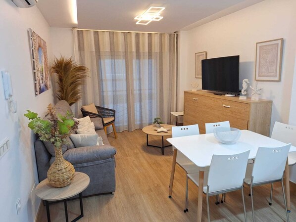Una sala de estar moderna con sofá, una mesa de comedor con sillas, televisión y un amplio ventanal con cortinas.