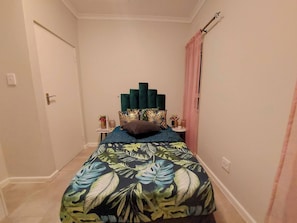 A narrow hallway leading to a bedroom with a bed featuring a tropical leaf pattern bedspread, a green upholstered headboard, and a small bedside table with a vase and a bottle. - Room-your Peaceful Haven, Unique Shared (Cape Town)