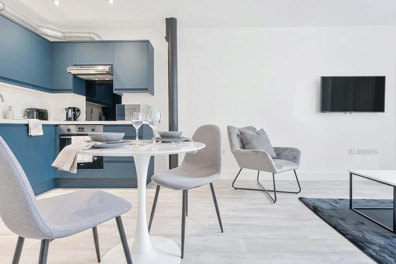A modern kitchen with blue cabinets, a dining table with chairs, and a flat-screen TV mounted on the wall.