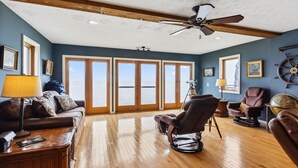 A spacious living room with a wooden floor, a ceiling fan, and large windows offering a view of the sea. - Lake Erie Getaway - Sunset Deck + Near GOTL Strip (Geneva)