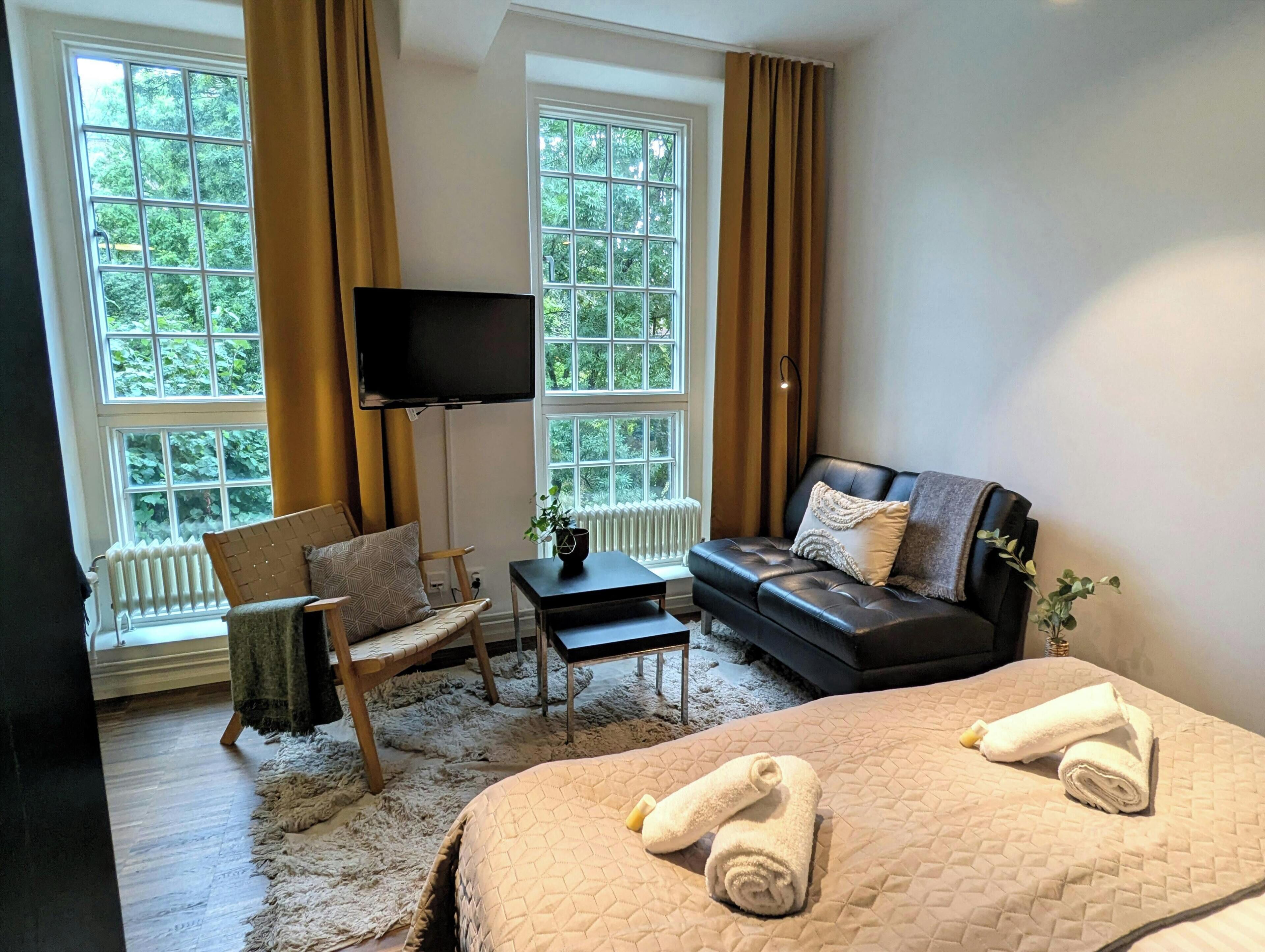 Hotellihuone, jossa on sänky, sohva, televisio, tuoli ja kaksi ikkunaa, joissa on verhot.