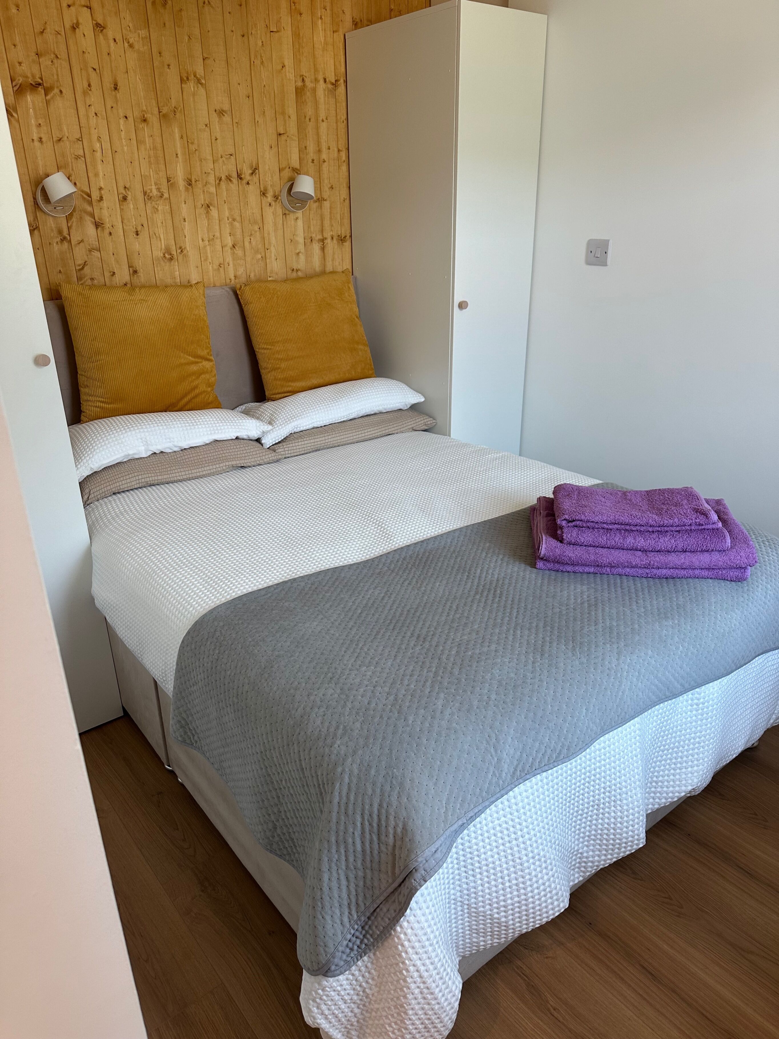 Un lit avec une couette grise et blanche, deux oreillers de couleur moutarde et une serviette violette posée sur le côté.