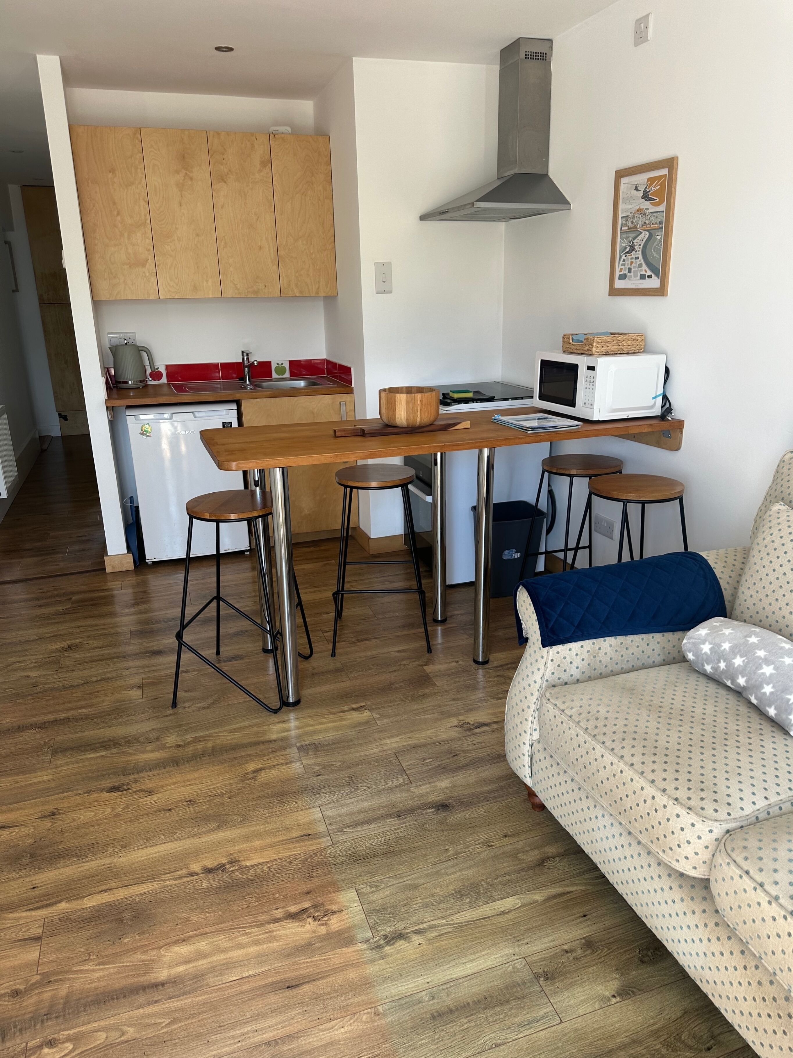A compact living space with a kitchenette, a dining table with stools, and a sofa with patterned cushions.