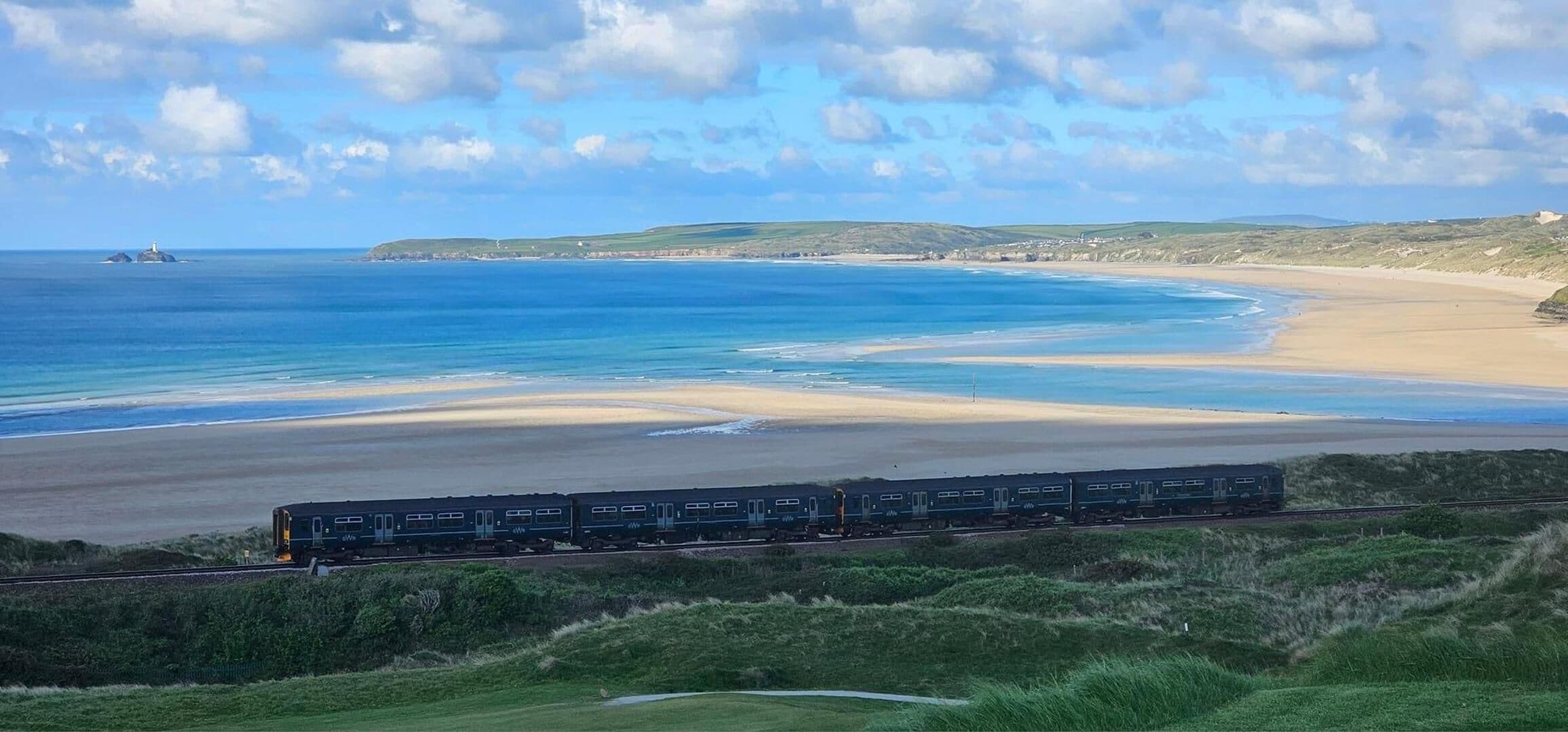 Un train circule sur une voie ferrée longeant la côte, avec une plage et l’océan en arrière-plan.