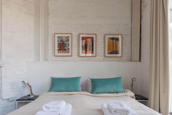 Habitación de hotel con una cama, dos almohadas, una mesita de noche, una lámpara y tres cuadros en la pared.