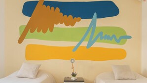 A room with two beds, a small table with a lamp, and a wall with abstract art.