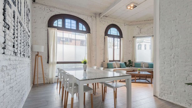 A spacious room with a white brick wall, a dining table with chairs, a sofa with cushions, and large windows with curtains.
