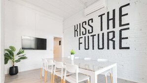 A modern meeting room with a white brick wall, a large TV mounted on the wall, a long table with white chairs, and a potted plant.
