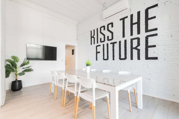 A modern meeting room with a white brick wall, a large TV mounted on the wall, a long table with white chairs, and a potted plant.