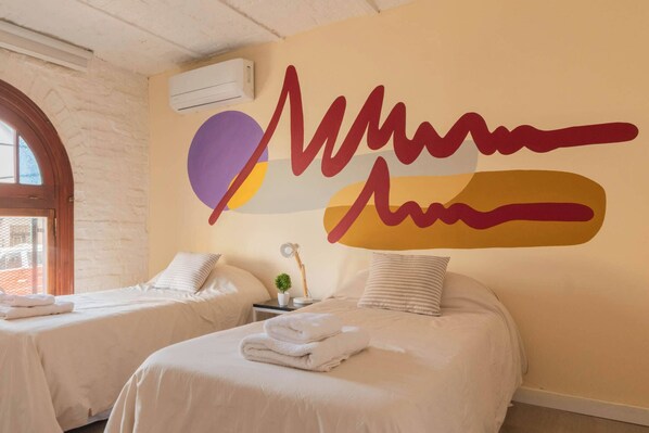 A hotel room with two beds, a wall with a large abstract design, a small table with a lamp, and a window with a view.