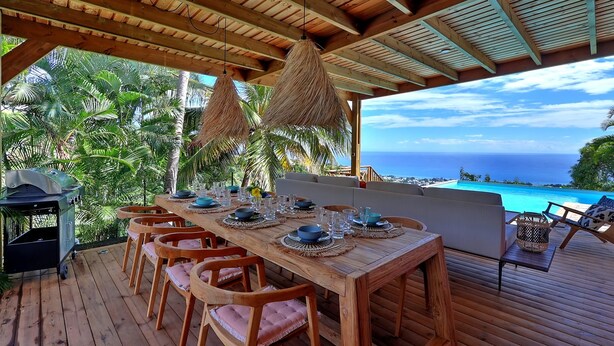 Una terraza de madera al aire libre con una mesa dispuesta para comer, rodeada de sillas, con vista a una playa y al océano.