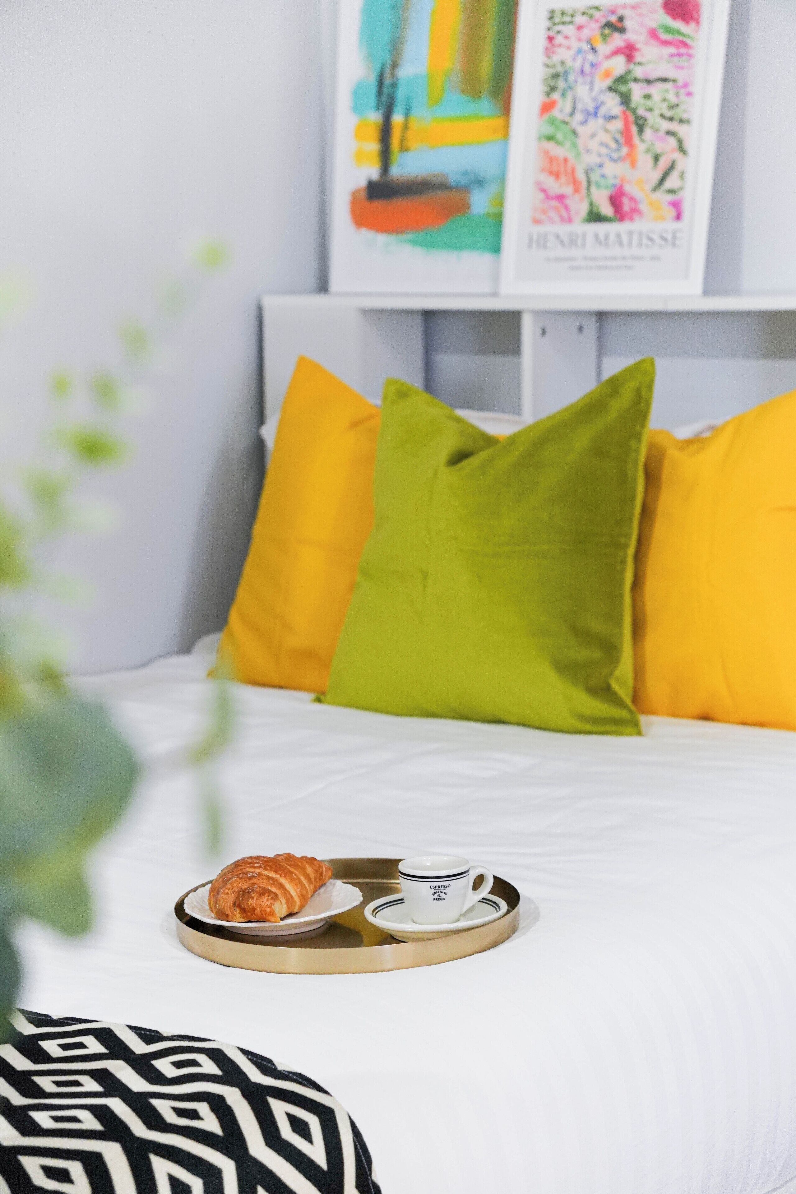 Una cama bien hecha con una bandeja de croissants y café, almohadas en tonos amarillo y verde, y una obra enmarcada en la pared.