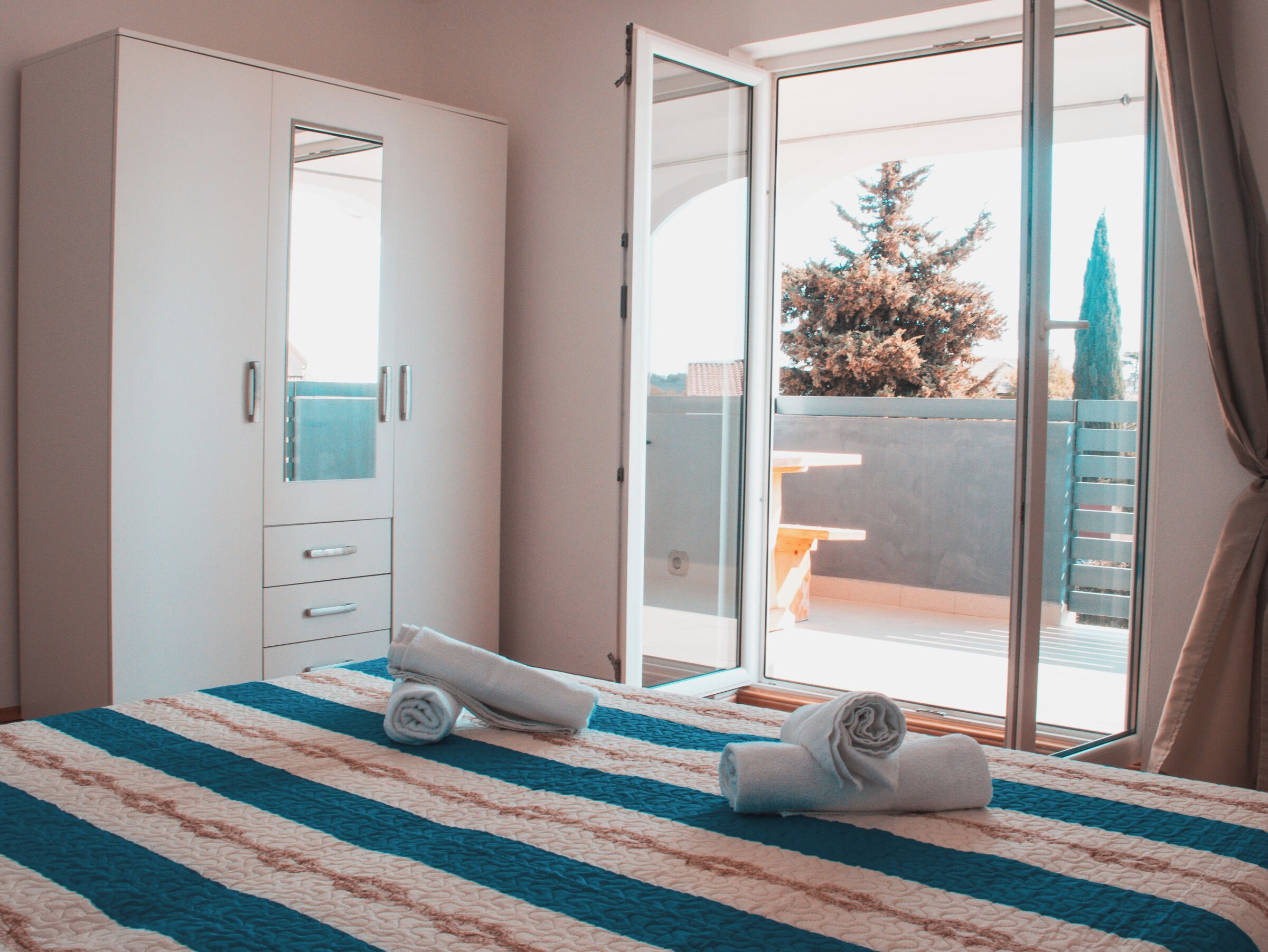 A bedroom with a bed, a wardrobe, and a sliding glass door leading to an outdoor area with a bench and a tree.