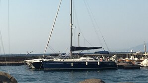 Un velero amarrado en un puerto deportivo, junto a otras embarcaciones, bajo un cielo despejado y con una cadena montañosa a lo lejos.