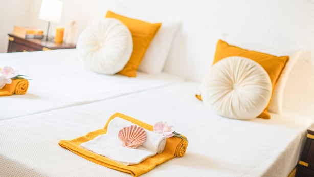 A neatly made bed with white and yellow bedding, a rolled towel, a seashell, and a pink flower. - Villa Dreams (Albufeira)