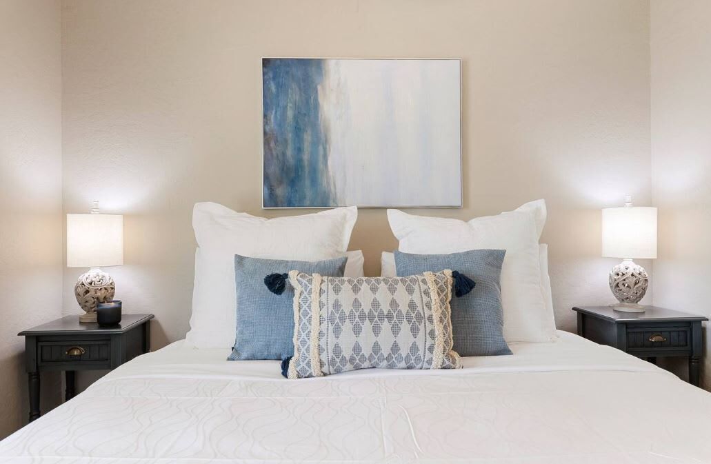 A neatly made bed with white bedding and decorative pillows, flanked by two bedside tables with lamps and decorative vases.