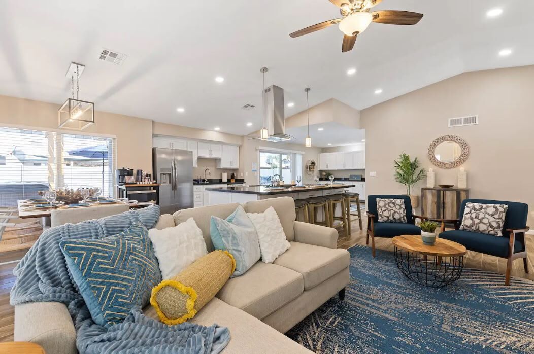 A modern living room with a sofa, coffee table, and two armchairs. There is a kitchen in the background with a refrigerator and a dining area.