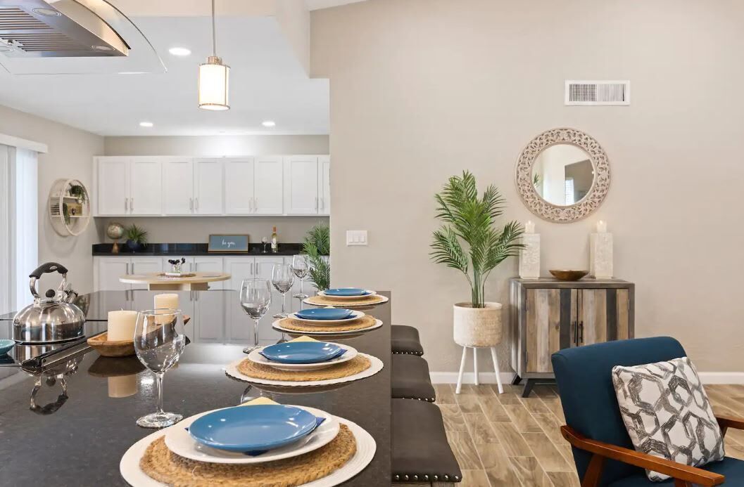 Une cuisine moderne avec des meubles blancs, un coin repas avec une longue table dressée pour six personnes, et un coin salon confortable avec un fauteuil bleu et un banc rembourré.