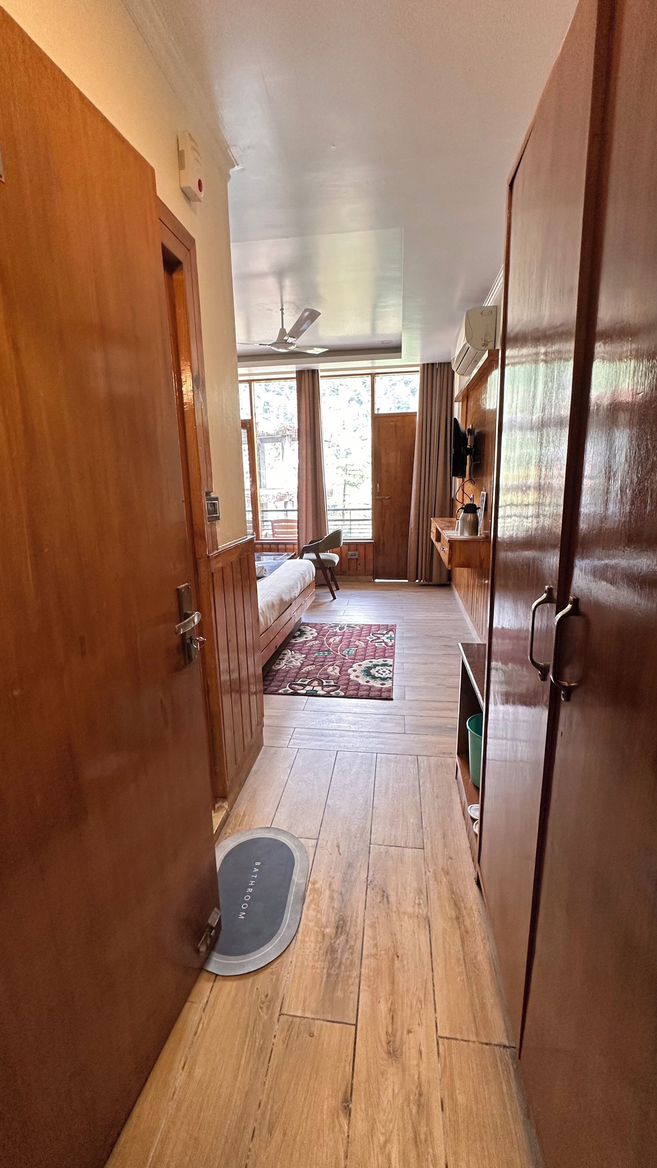 A hallway leading to a bedroom with a bed, a desk, and a chair. There is a bathroom with a mat labeled 'BATHROOM' and a ceiling fan.