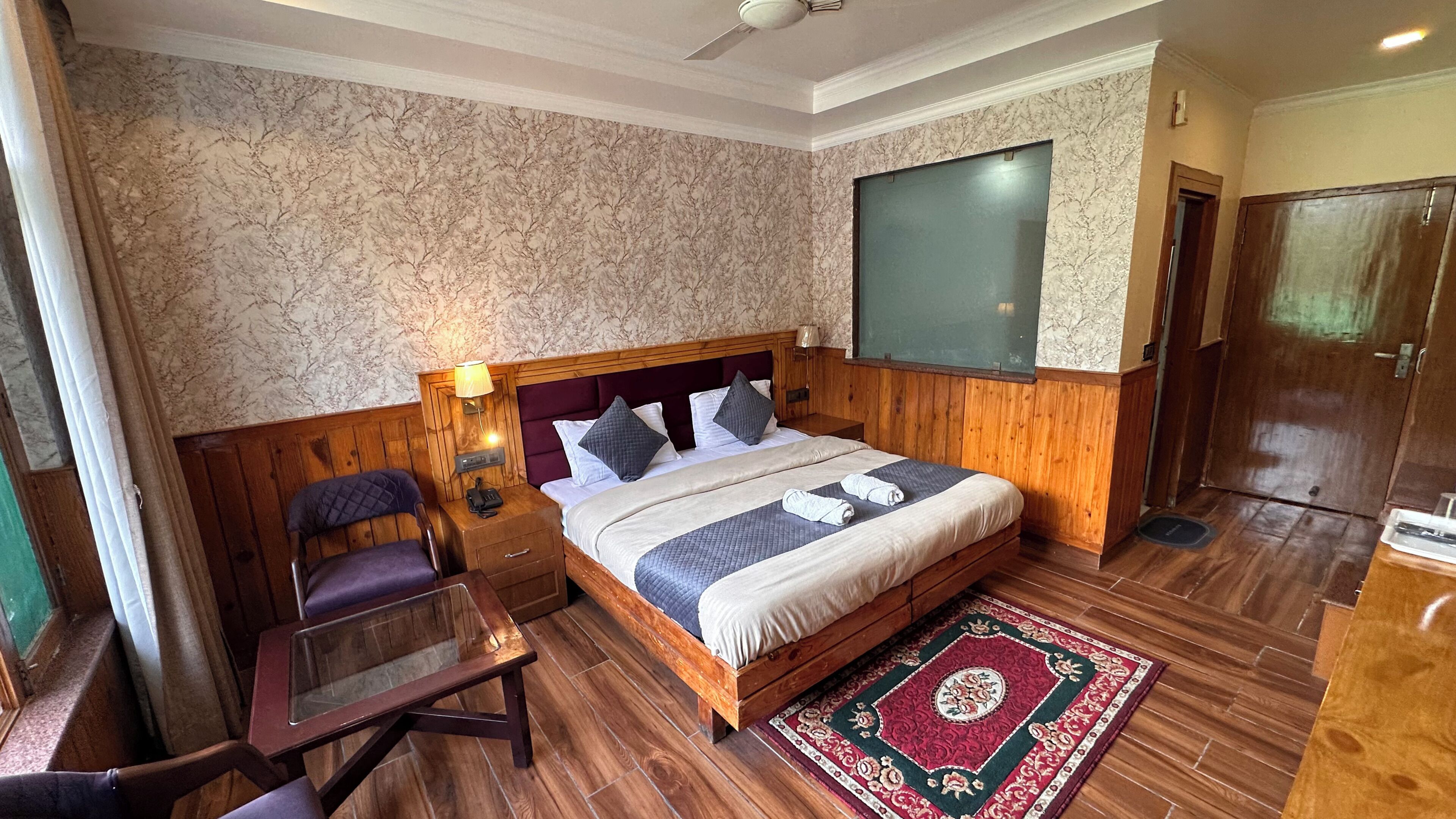 A hotel room with a wooden bed, a nightstand, a chair, a TV, and a wooden wardrobe.