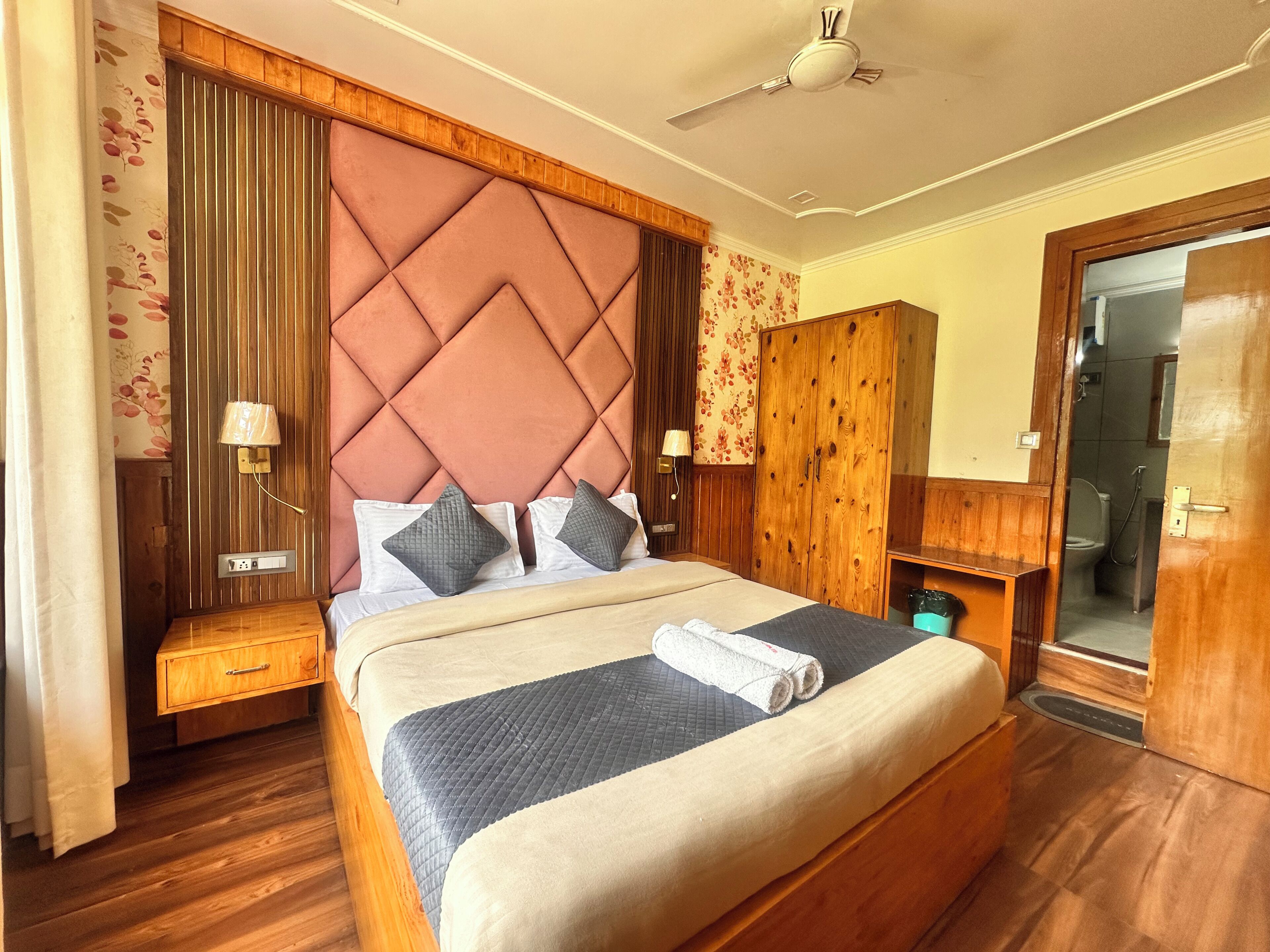 Habitación de hotel con una cama grande, cabecera de madera, mesitas de noche, un armario y un baño visible a través de una puerta abierta.