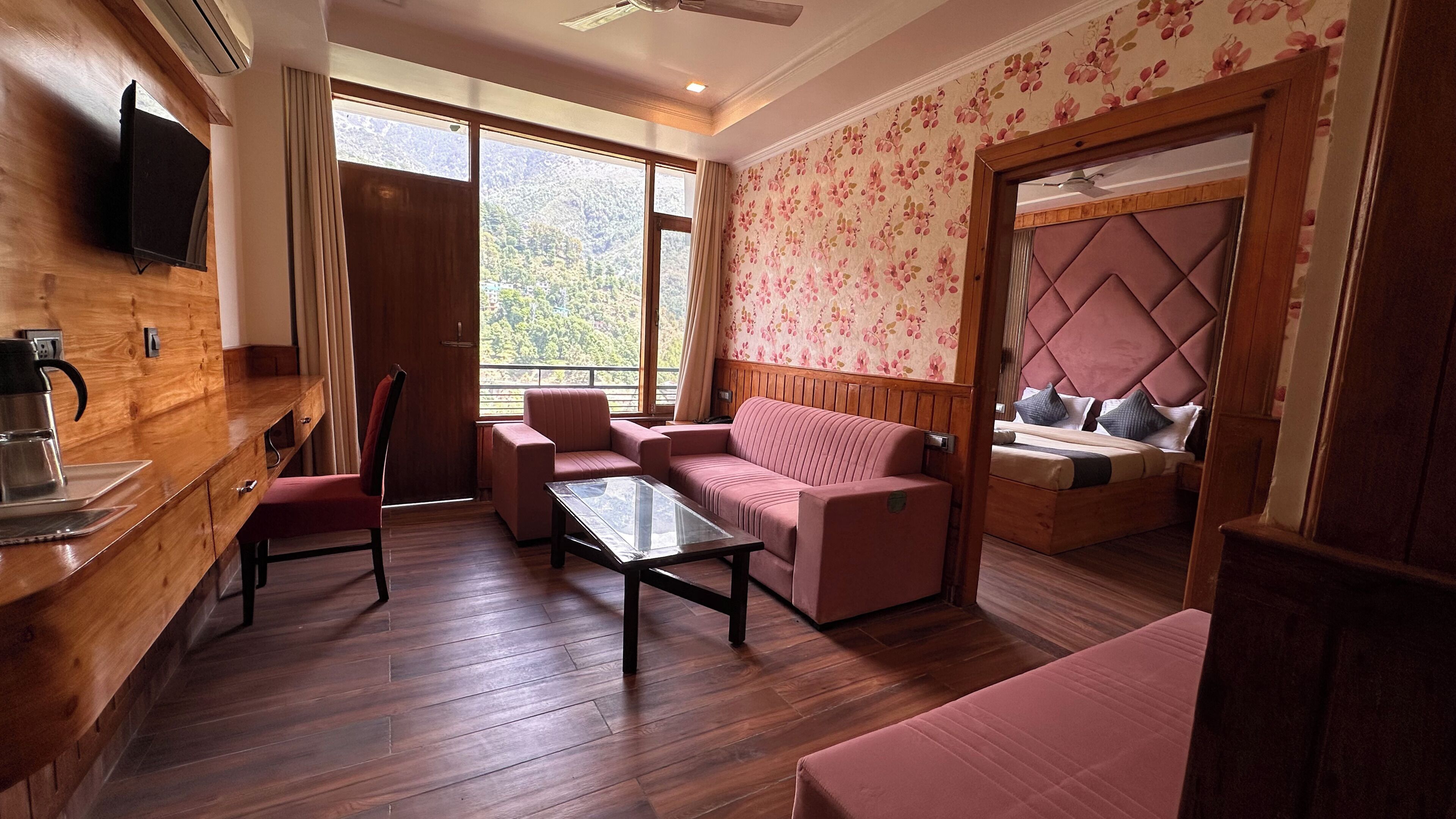 A room with a wooden floor, a sofa, a chair, a table, a bed with a tufted headboard, and a television mounted on the wall.