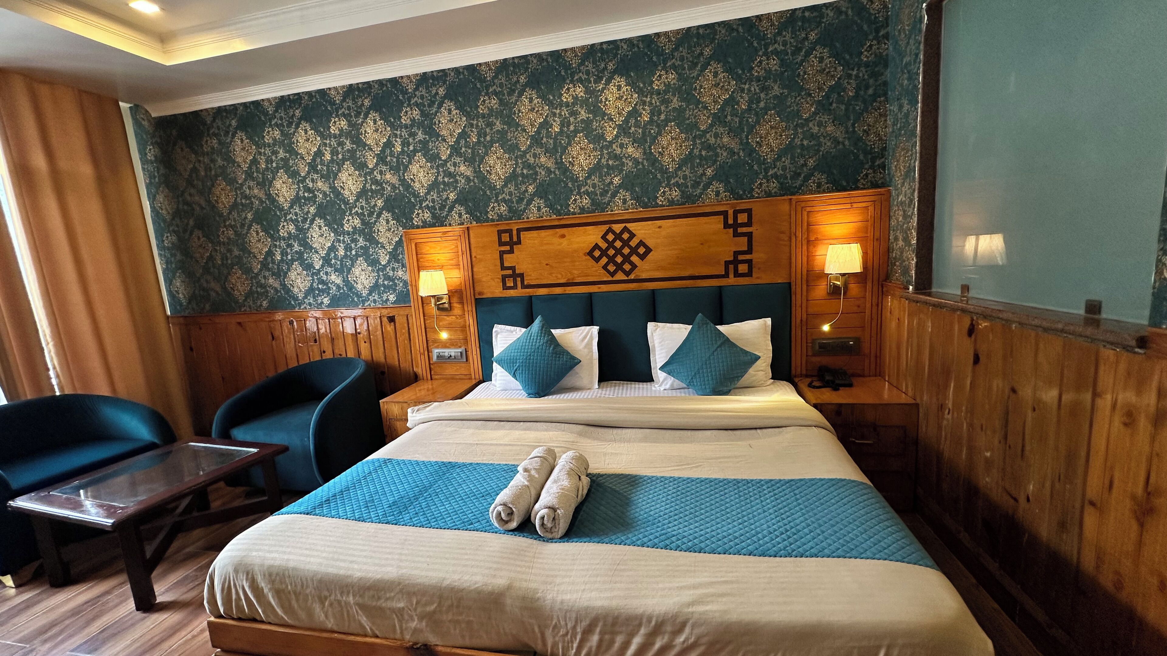 Ein Hotelzimmer mit einem Bett, zwei Stühlen, einem Holz-Kopfteil, einem Nachttisch und einer Wand mit strukturiertem Wandpapier.