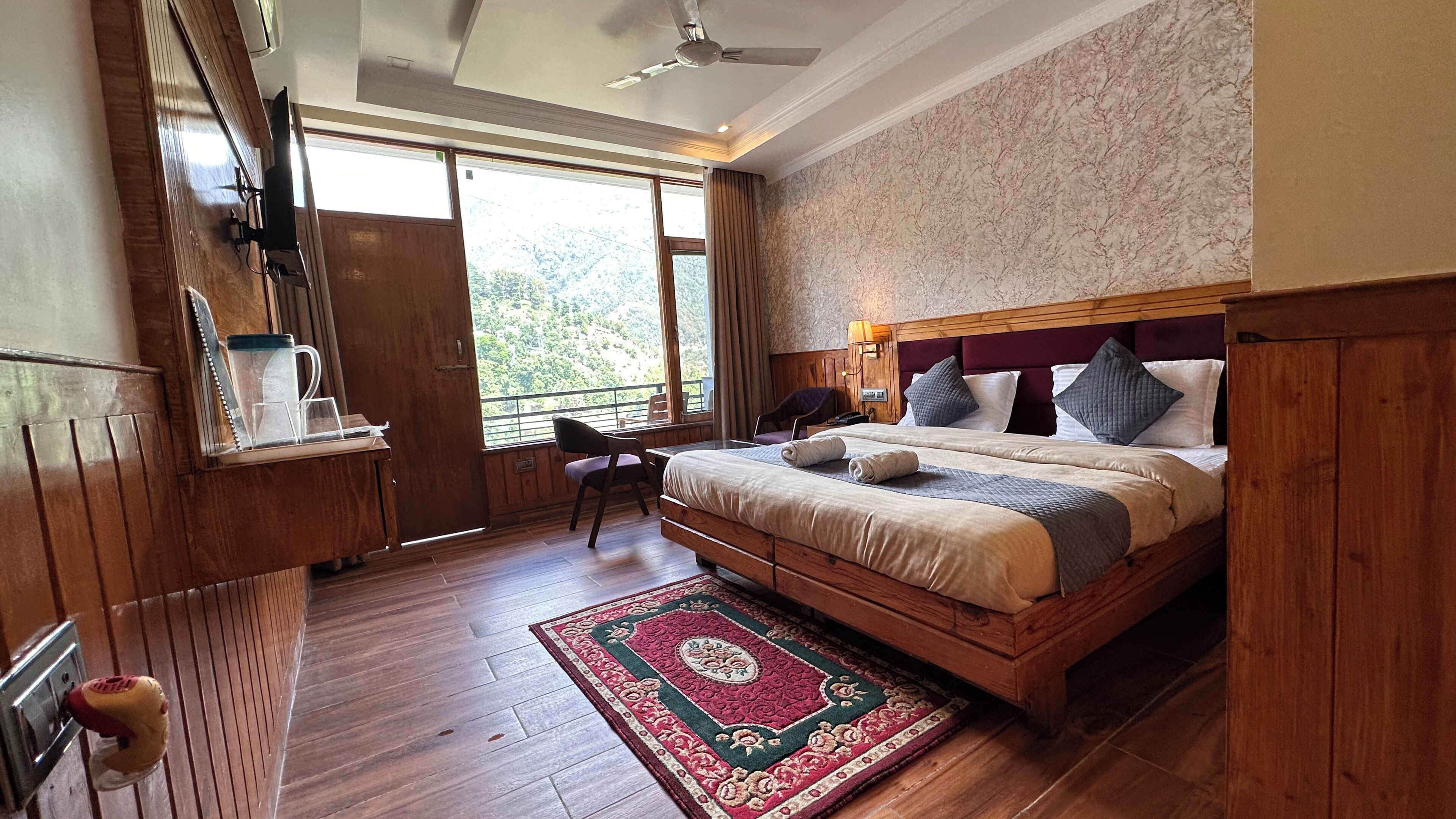A bedroom with a large bed, wooden headboard, a desk with a chair, a window with curtains, and a ceiling fan.