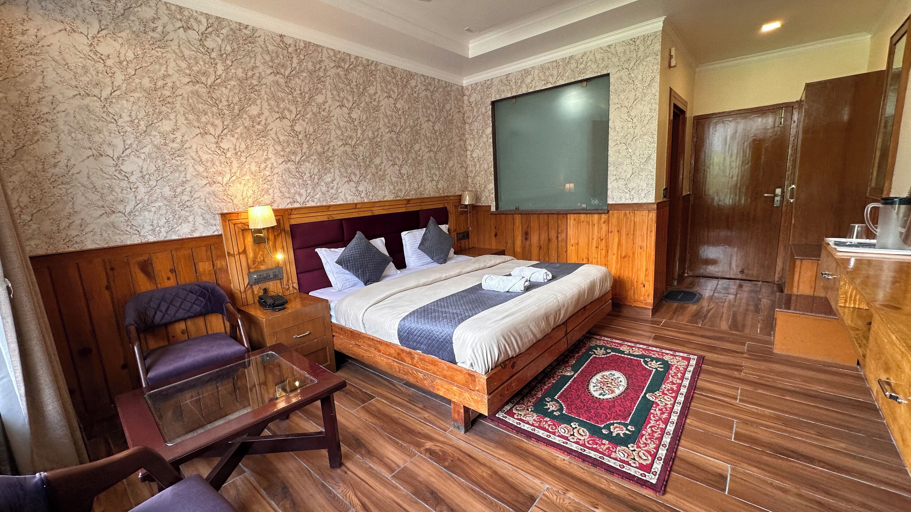 A hotel room with a wooden bed, a nightstand with a lamp, a wooden chair, a glass table, and a large mirror.