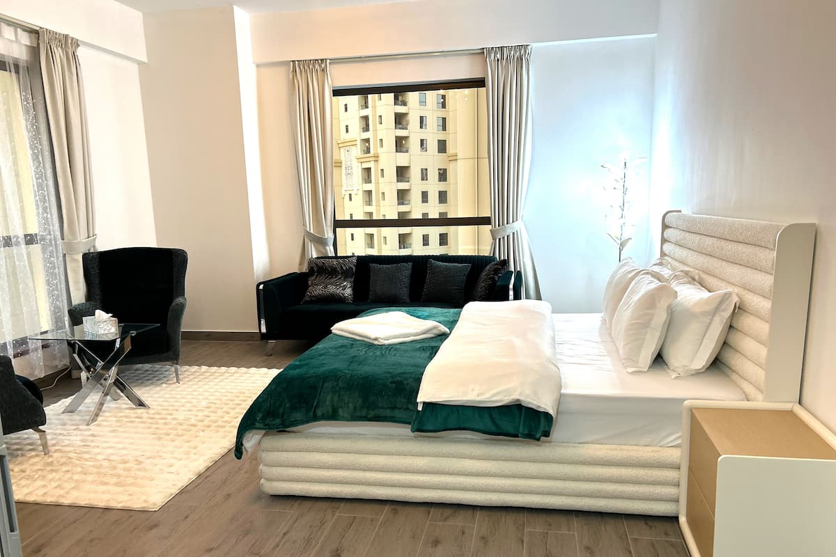 Une chambre d’hôtel moderne dotée d’un grand lit, d’un coin salon avec un fauteuil et une table, et offrant une vue sur un immeuble par la fenêtre.