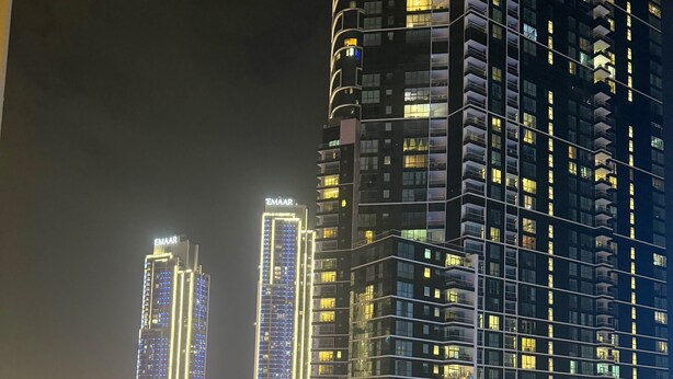 Un edificio alto con ventanas y balcones iluminados, un área de piscina con diseño hundido, y una calle concurrida con vehículos.