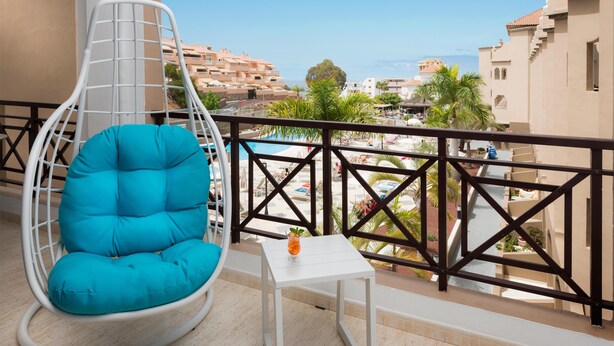 A balcony with a turquoise hanging chair, a small white table with a drink, and a view of a pool and buildings. - Saint George Apartamentos      (Arona)