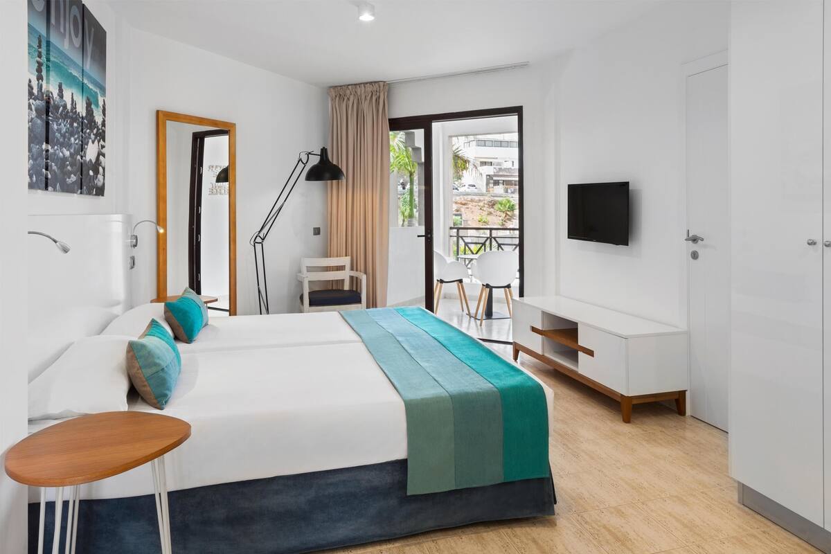 A modern hotel room with a bed, a desk, a chair, a TV, and a balcony with a view of the city.