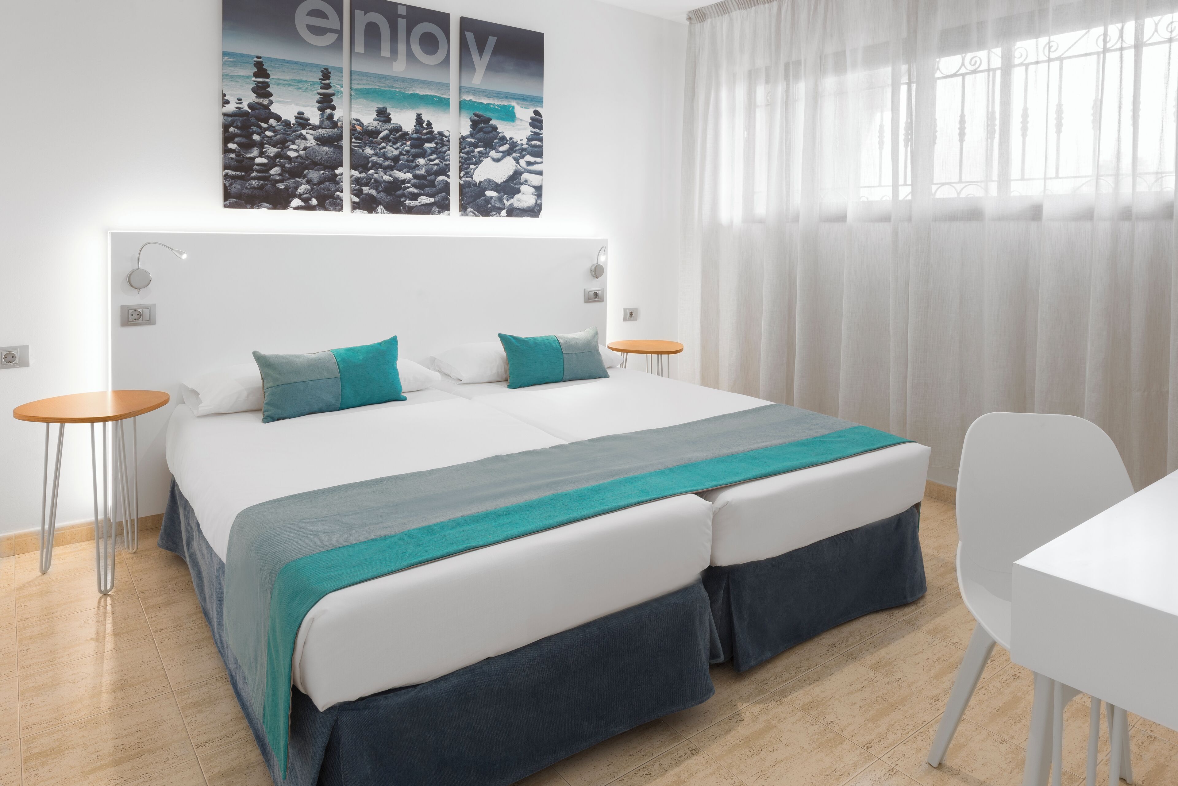 A modern hotel room with a large bed, two bedside tables, a small round table, and a view of the ocean through sheer curtains.