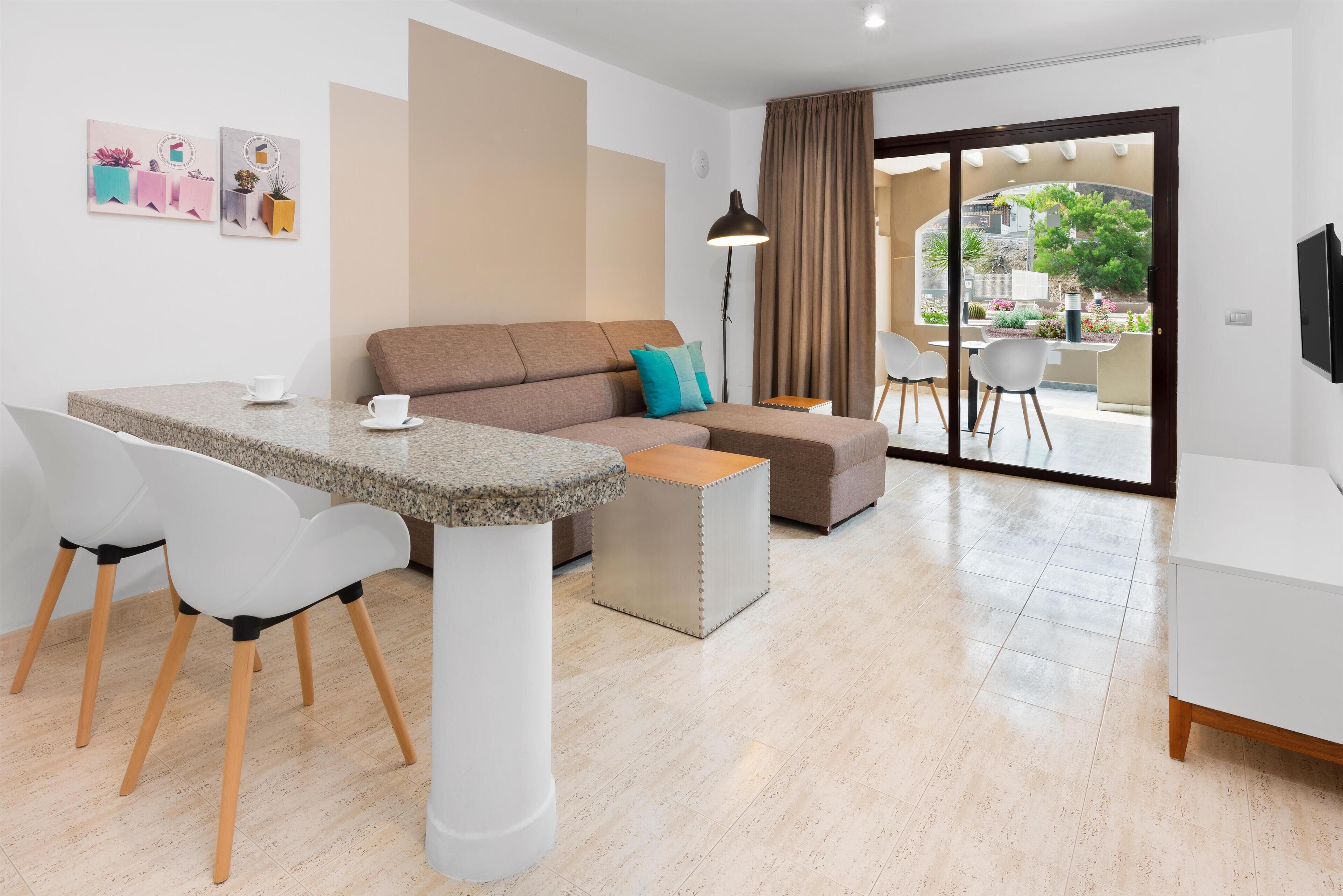 Ein modernes Wohnzimmer mit einer Couch, einem Esstisch mit Stühlen, einem Couchtisch und einem großen Fenster mit Vorhängen.