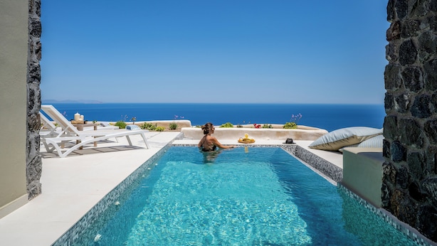 Una piscina con vista al mar, rodeada de muros de piedra y sillones blancos para tomar el sol.