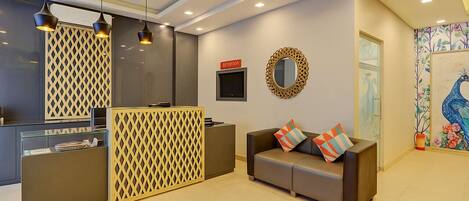 A reception area with a reception desk, a waiting area with a sofa and cushions, a decorative mirror, and a wall-mounted TV.