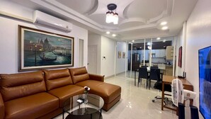 A modern living room with a brown leather sofa, a glass coffee table, a painting of a canal scene, and a view into a dining area with a kitchen. - Valley 40 - 2 bdr Private Villa Near Phuket Town (Kathu)