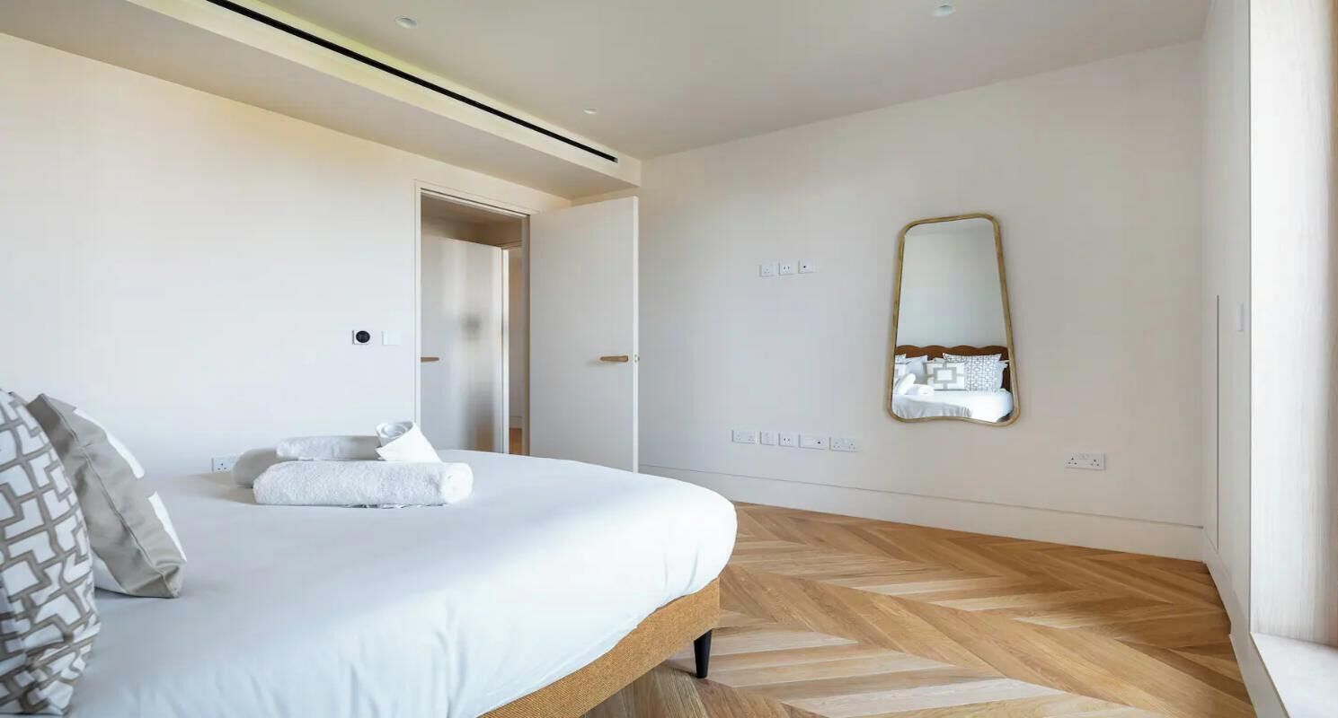 Une chambre moderne avec un grand lit, un parquet en bois disposé en motif chevrons, un miroir et une porte donnant sur une autre pièce.