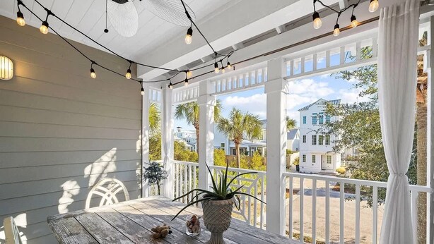 A covered porch with a wooden table, string lights, and a view of houses and palm trees.