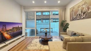A modern living room with a flat-screen TV, a beige sofa, a coffee table, and a large window overlooking a building.