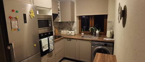 A compact kitchen with a stainless steel refrigerator, microwave, oven, and dishwasher. There is a wooden countertop, a sink, and a window above the sink.