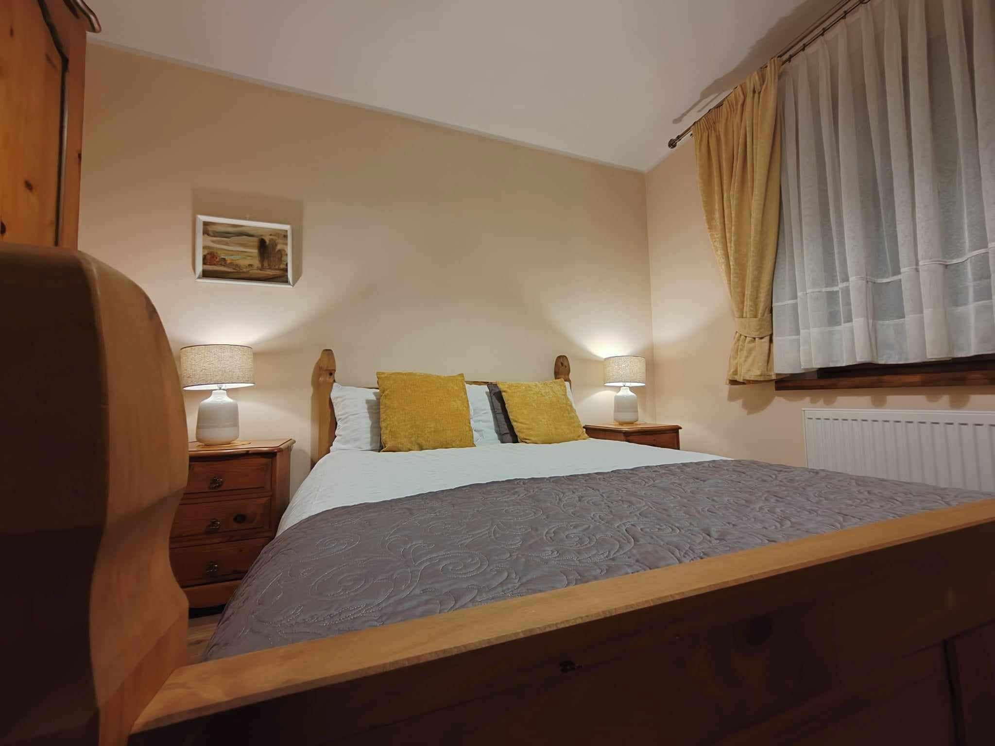 A bedroom with a wooden bed frame, two bedside tables with lamps, a radiator, and a window with curtains.