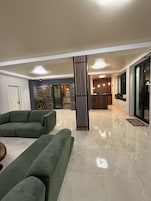 Una sala moderna con un sofá modular, un columnario decorativo alto y una cocina al fondo.