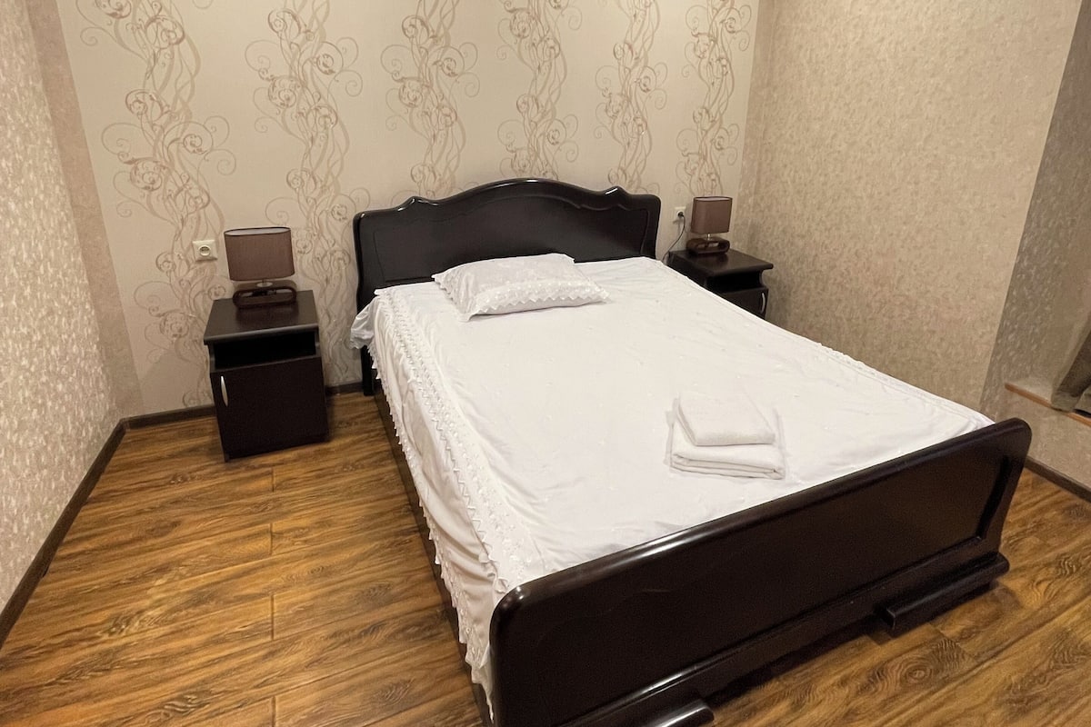 Ein ordentlich bezogenes Bett mit weißen Leinen und einem gefalteten Handtuch, ein Holzboden und eine Wand mit floralem Muster.