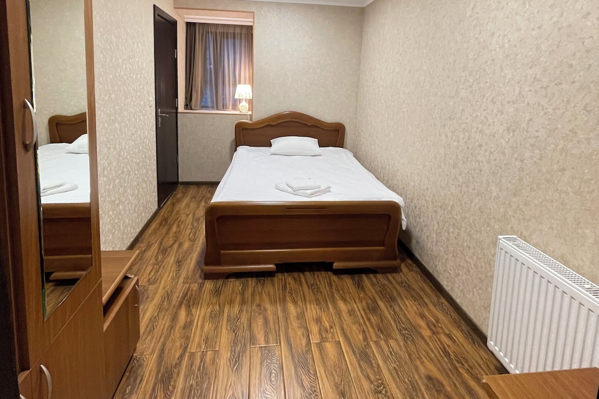Ein Schlafzimmer mit Holzboden, einem Bett mit weißer Bettwäsche, einem Fenster mit Vorhängen, einem Heizkörper und einem Spiegel an der Tür.