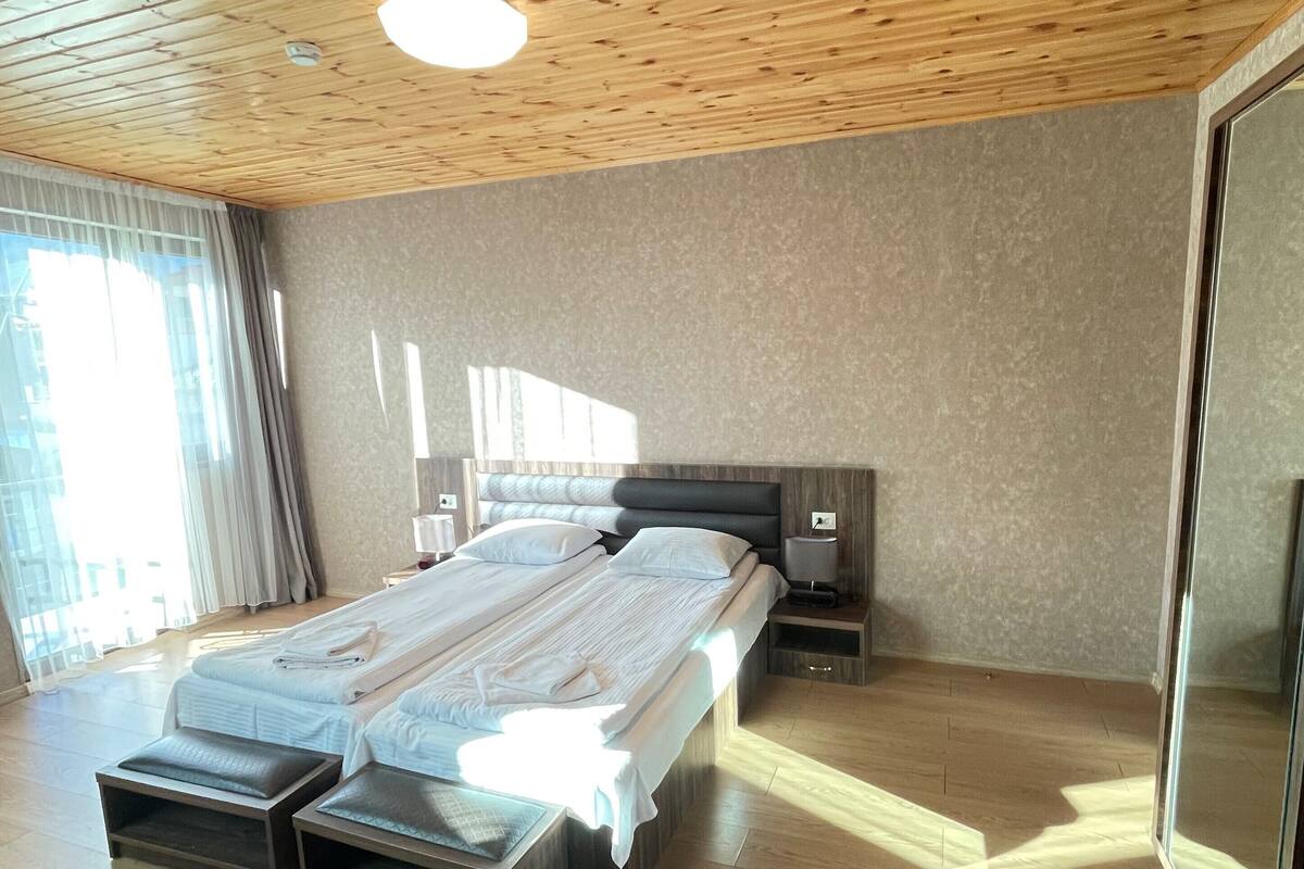 Ein modernes Schlafzimmer mit Holzbalkendecke, einem großen Bett mit Kopfteil, Nachttischen und einem großen Fenster mit Vorhängen.