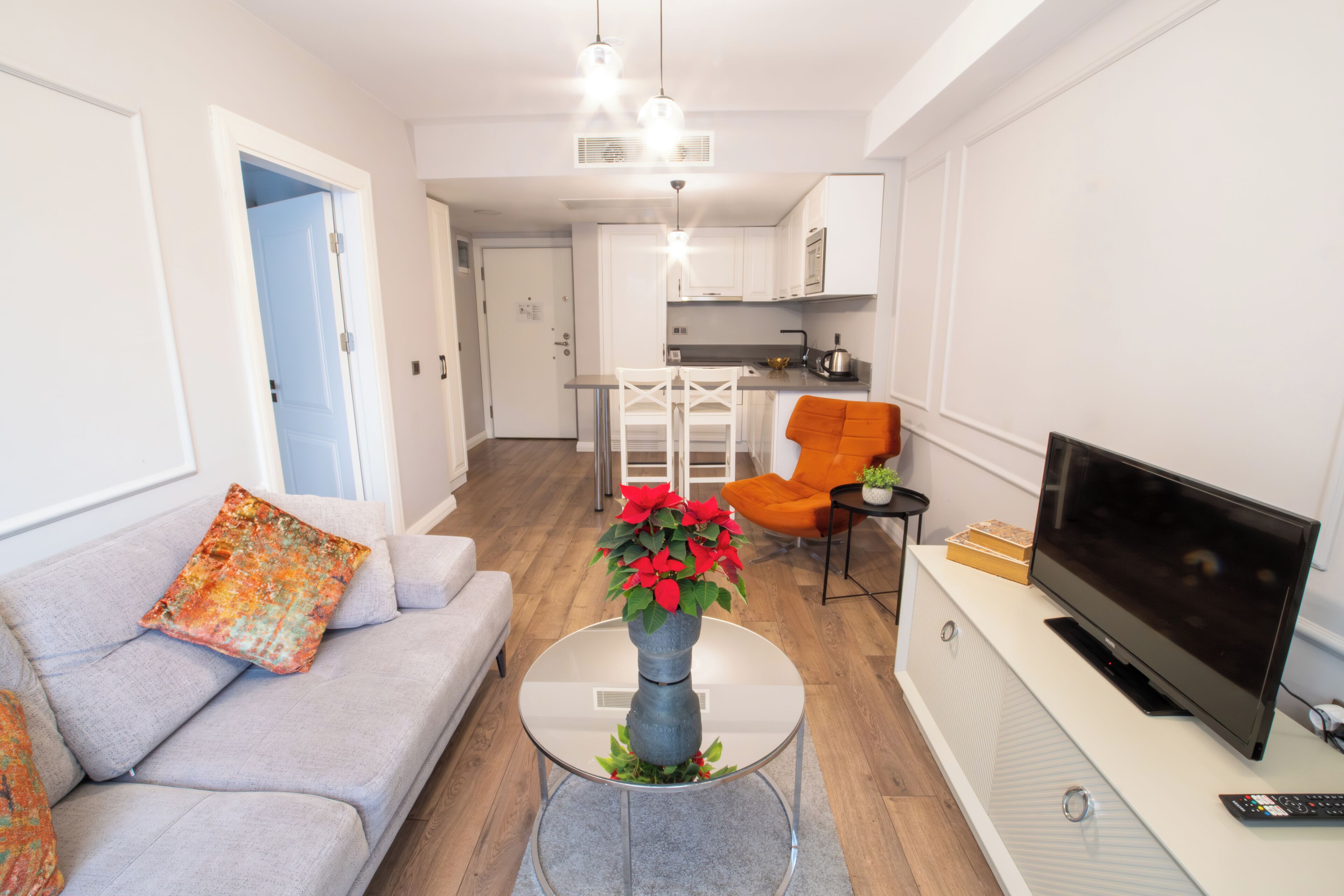 Een moderne woonkamer met een bank, een bijzettafeltje met een vaas, een televisie en een keuken op de achtergrond.